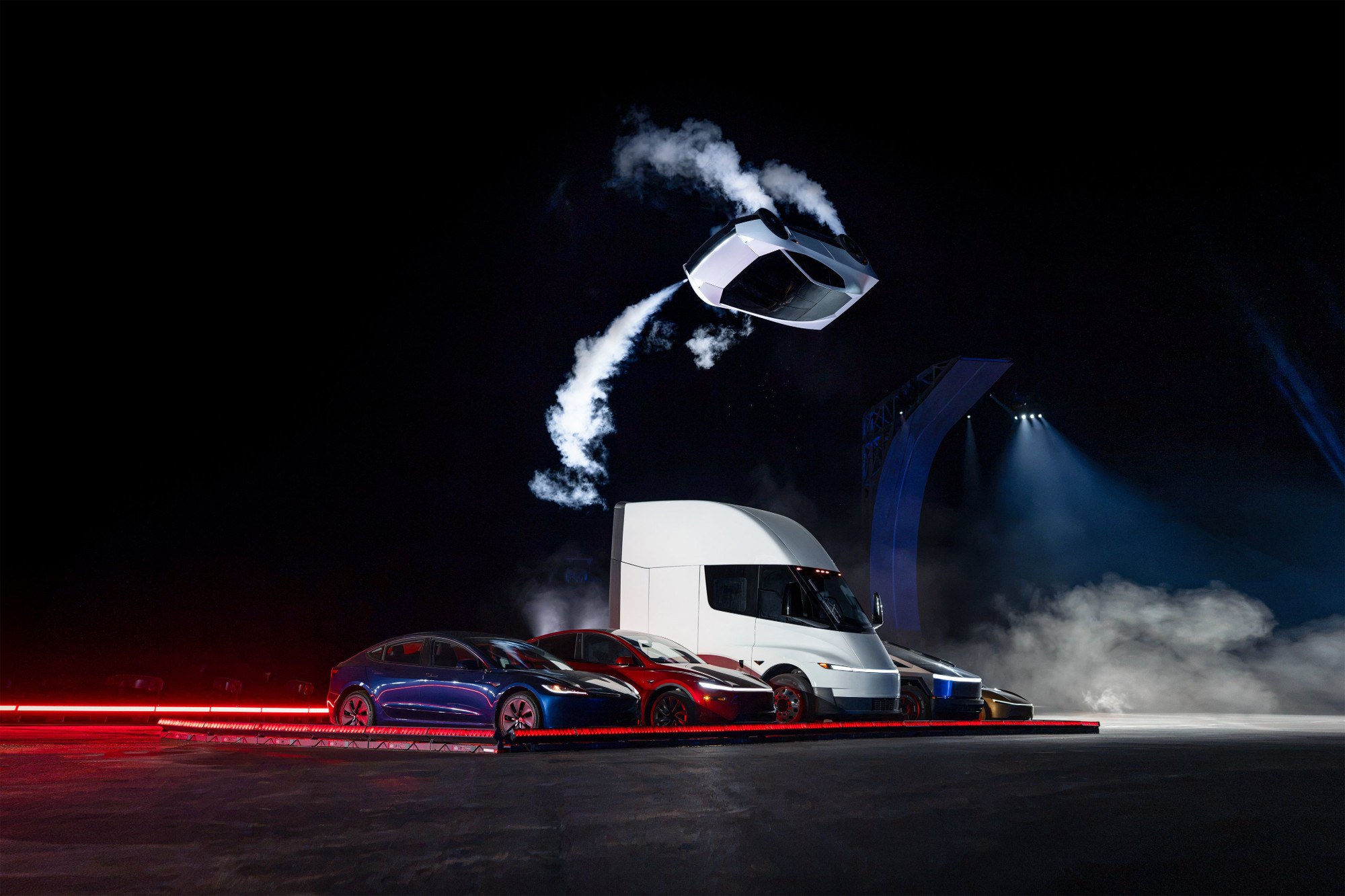 A white Tesla Roadster airborne and tilted in a dark arena, trailing white thruster smoke, above a row of Tesla vehicles: a blue Model 3, a red Model Y, a white Semi truck and a Cybertruck, lit by red floor lights.